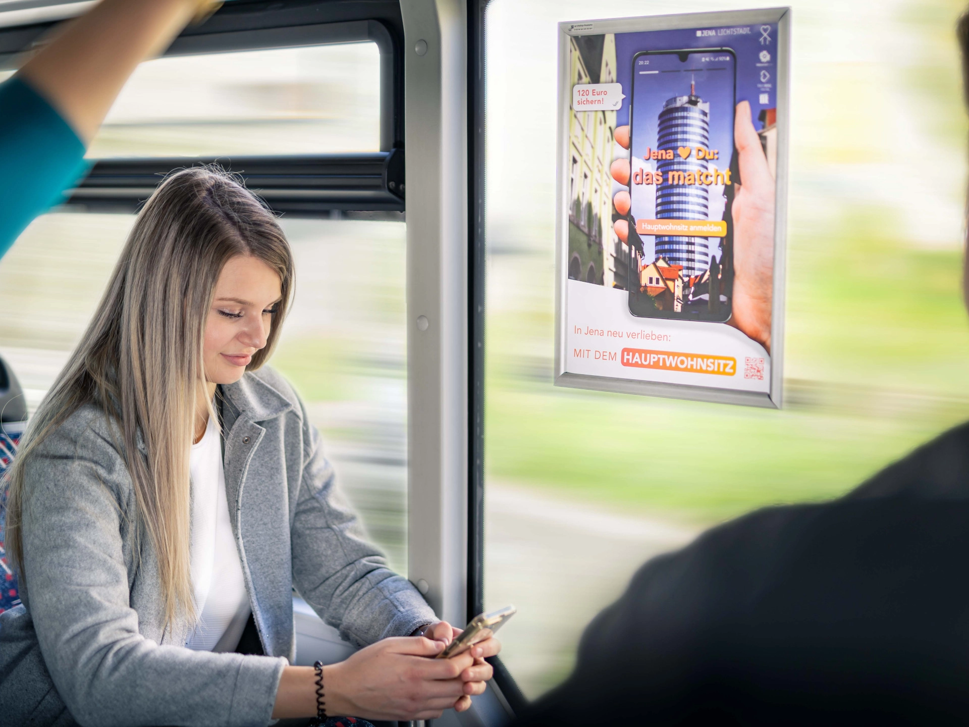 Eine Frau sitzt mit Handy im Bus und neben ihr ist Plakatwerbung zu sehen.