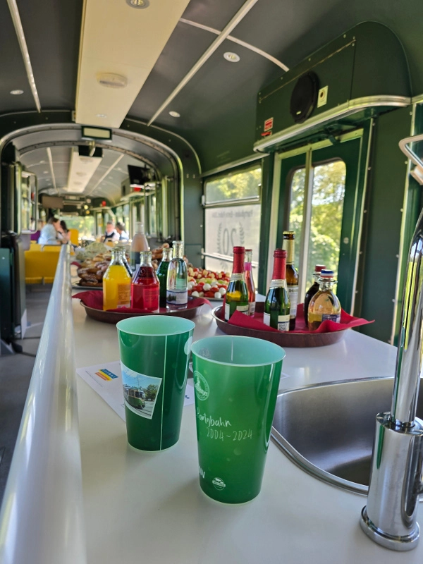 Im Vordergrund sieht man Eventbecher und Buffet auf dem Tresen stehen, im Hintergrund sind Fahrgäste.